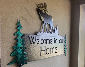 May include: Metal sign with a welcome to our home message. The sign features a silhouette of an elk with trees on either side.