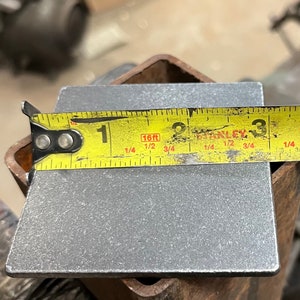 May include: A close-up of a metal square plate being measured with a yellow tape measure. The tape measure reads "1 1/4 16ft 1/2 3/4".