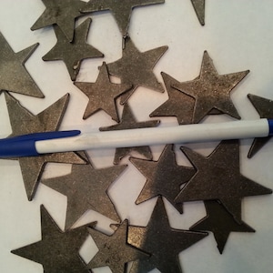 May include: A pile of metal star shapes in a silver color. The stars are scattered on a white surface with a blue and white pen lying on top of them.