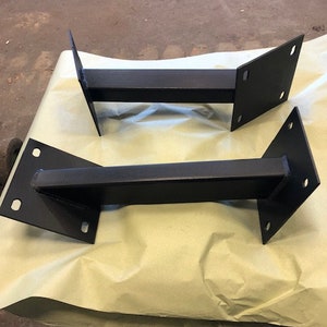 May include: Two black metal brackets with a rectangular base and a long, straight bar connecting the two sides. The brackets are on a light brown surface.