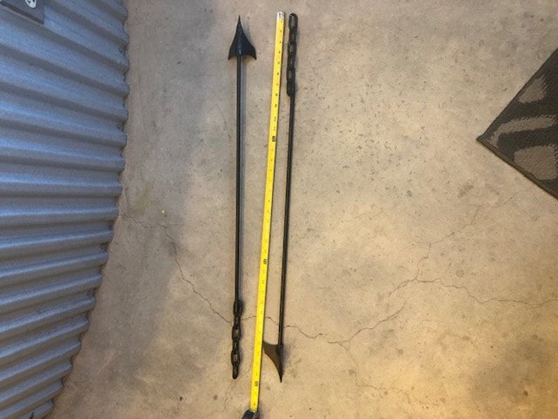 May include: Two black metal rods with pointed ends. One rod has a chain attached to it. The rods are lying on a concrete floor.