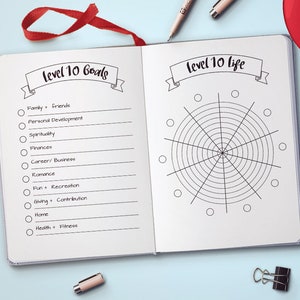 Puede incluir: Un diario abierto con los textos "Level 10 Goals" y "Level 10 Life". La p&aacute;gina izquierda enumera objetivos como familia, finanzas y salud. La p&aacute;gina derecha presenta un diagrama circular. Una cinta roja como marcador es visible.