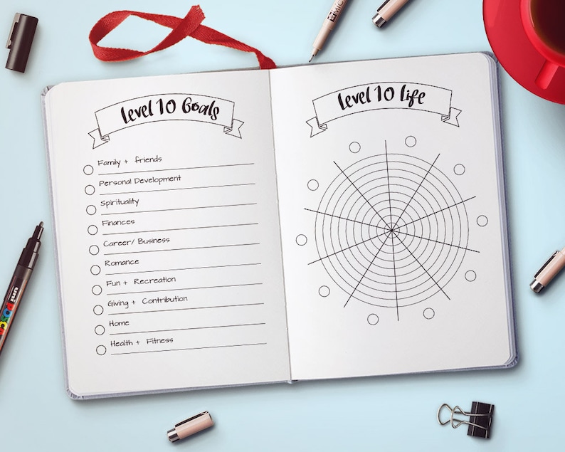 Puede incluir: Un diario abierto con los textos "Level 10 Goals" y "Level 10 Life". La p&aacute;gina izquierda enumera objetivos como familia, finanzas y salud. La p&aacute;gina derecha presenta un diagrama circular. Una cinta roja como marcador es visible.