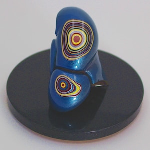May include: A blue abstract sculpture with concentric circles in yellow, red, and white. The sculpture sits on a dark gray circular base. The artwork has a glossy finish and a unique, eye-catching design.