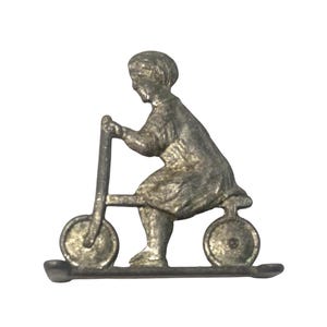 Puede incluir: Un broche de metal plateado que representa a una persona montada en un patinete vintage. La figura está sentada en el patinete con las manos en el manillar. El patinete tiene dos ruedas y un reposapiés. El broche es una representación detallada en miniatura.