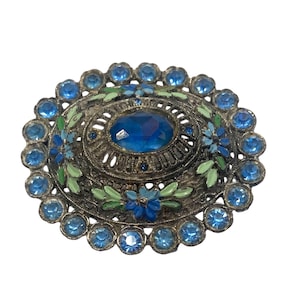 Op de afbeelding: Ovale vintage broche met een centrale blauwe edelsteen omringd door kleinere blauwe stenen. De zilverkleurige metalen rand heeft blauwe en groene emaille bloemdetails, wat een klassiek, elegant ontwerp creëert.