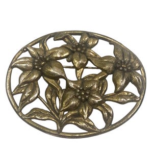 Puede incluir: Broche ovalado de estilo antiguo con diseño floral. El broche presenta cinco flores detalladas con hojas, engastadas en un marco calado. El metal tiene un aspecto deslustrado y envejecido.