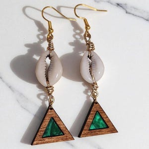 May include: A pair of dangle earrings featuring a natural seashell, gold-colored wire, and a wooden triangle with a green resin inlay. The earrings have gold-colored ear wires and are displayed on a white marble surface.