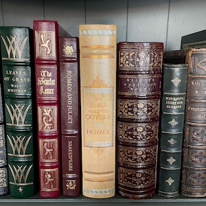 May include: A selection of antique books with diverse colours and designs. Titles include "Leaves of Grass", "The Scarlet Letter", "Romeo and Juliet", "The Iliad and the Odyssey", and "Kipling Lysed Der Sluktes". The books are displayed on a shelf.