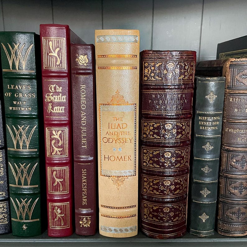 May include: A selection of antique books with diverse colours and designs. Titles include "Leaves of Grass", "The Scarlet Letter", "Romeo and Juliet", "The Iliad and the Odyssey", and "Kipling Lysed Der Sluktes". The books are displayed on a shelf.