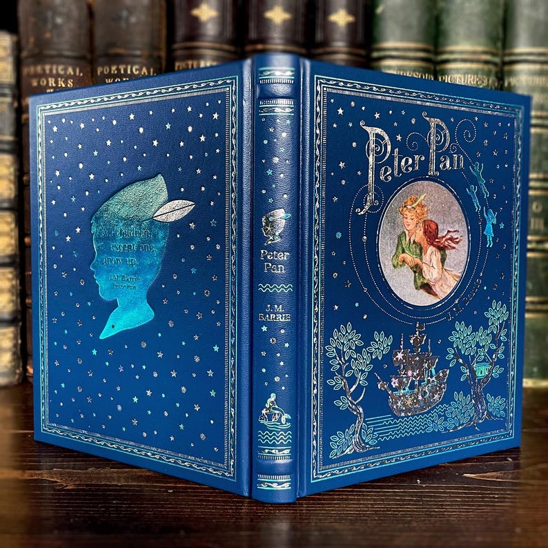 May include: A hardcover book titled "Peter Pan" by J.M. Barrie. The book has a blue cover with silver foil accents, including stars and illustrations of Peter Pan and other characters. The spine features the title and author.