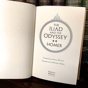 May include: An open book displaying the title "The Iliad and the Odyssey" by Homer. The text is within a circular design with decorative borders. Published by Barnes & Noble, New York.