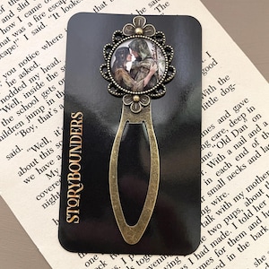 May include: A black bookmark with a bronze-colored metal design. The top features a round image of a couple embracing, framed by a floral design. The text "STORYBOUNDERS" is printed vertically on the bookmark.