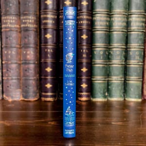 May include: A blue book spine with the title "Peter Pan" and author J.M. Barrie. The spine features a starry design with illustrations of Peter Pan and a ship. The book is set against a backdrop of antique books.