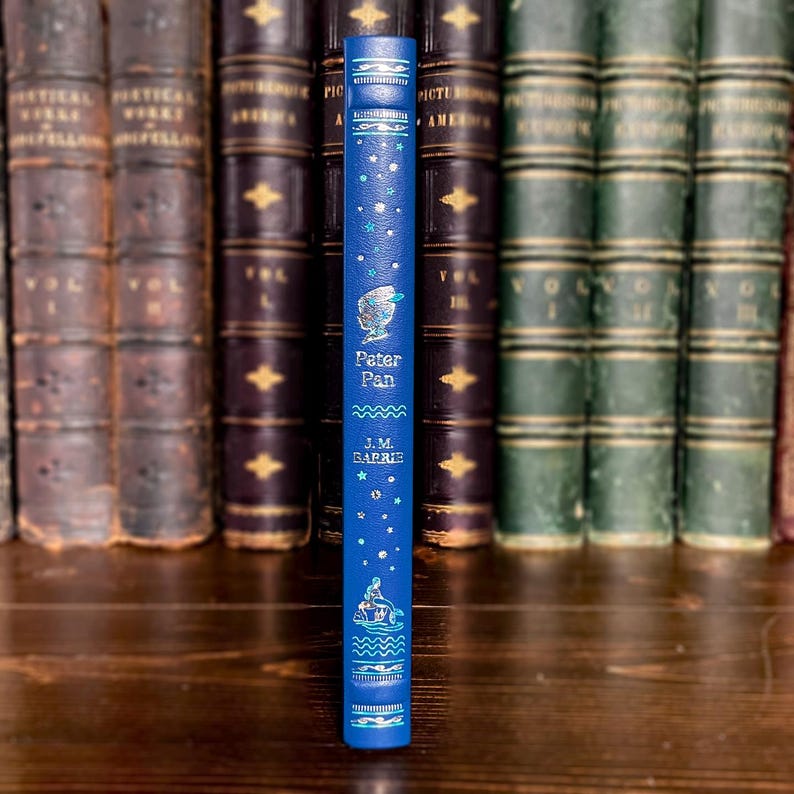 May include: A blue book spine with the title "Peter Pan" and author J.M. Barrie. The spine features a starry design with illustrations of Peter Pan and a ship. The book is set against a backdrop of antique books.