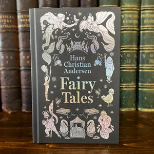 May include: A hardcover book titled "Fairy Tales" by Hans Christian Andersen. The cover is dark gray with iridescent illustrations of mermaids, a castle, and other fairy tale characters. The title is in a shimmering, metallic font.