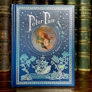 May include: A hardcover book titled "Peter Pan" with a dark blue cover. The cover features an illustration of Peter Pan and Wendy, a ship, and fairies. The author's name, J.M. Barrie, is printed at the bottom. The book is surrounded by other books.