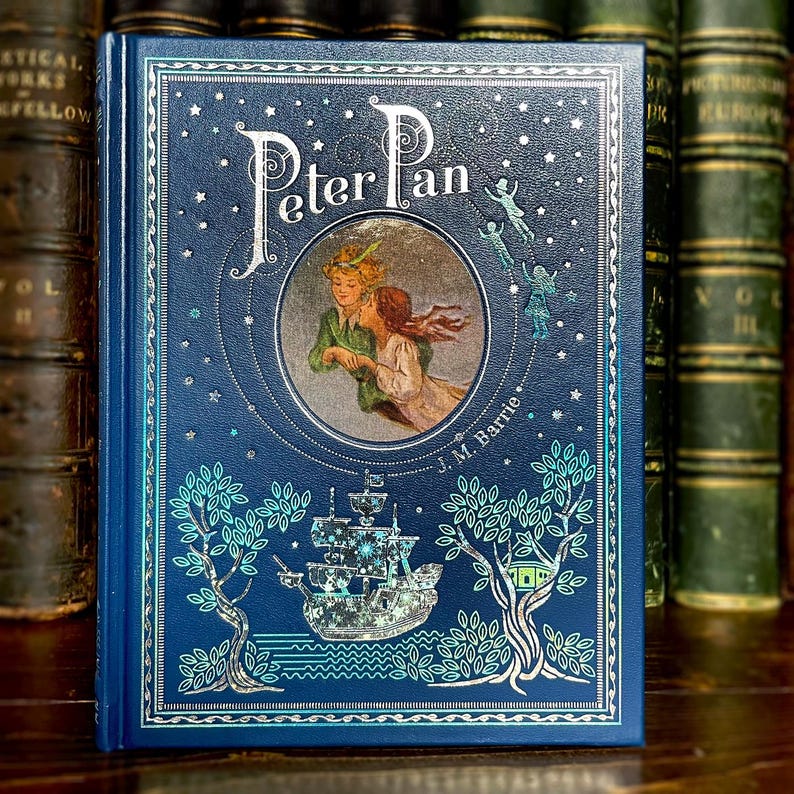May include: A hardcover book titled "Peter Pan" with a dark blue cover. The cover features an illustration of Peter Pan and Wendy, a ship, and fairies. The author's name, J.M. Barrie, is printed at the bottom. The book is surrounded by other books.