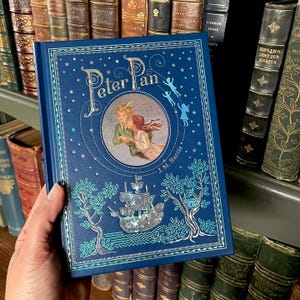 May include: A hardcover book titled "Peter Pan" with a navy blue cover. The cover features an illustration of Peter Pan and Wendy, a ship, and decorative trees. The title is in a stylized font, and the author's name, J.M. Barrie, is printed below the illustration.