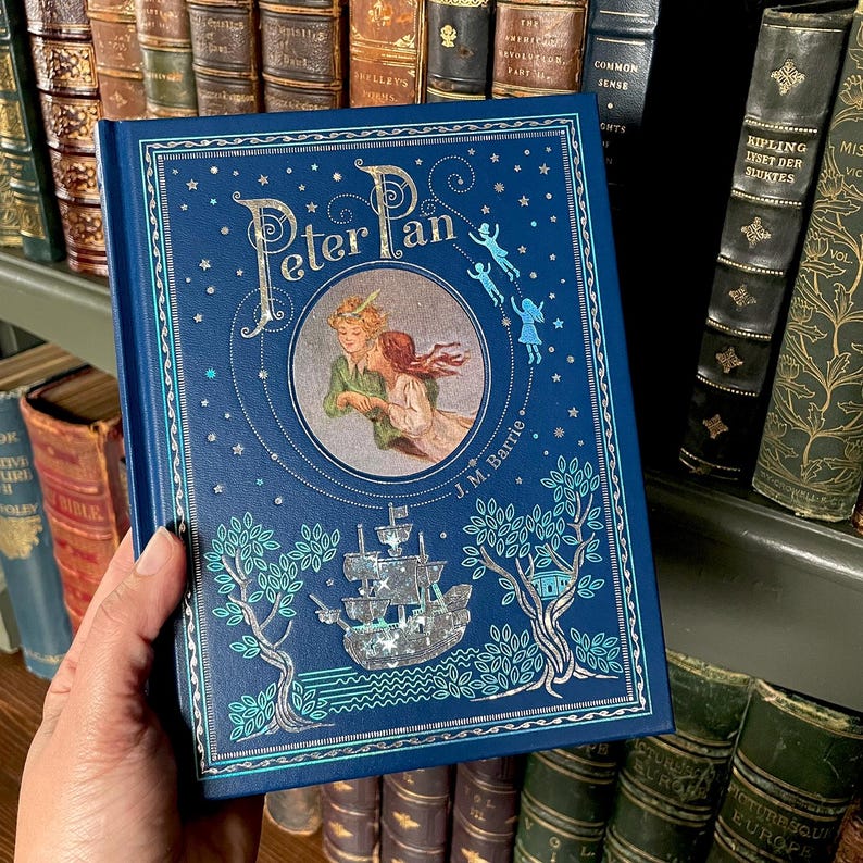 May include: A hardcover book titled "Peter Pan" with a navy blue cover. The cover features an illustration of Peter Pan and Wendy, a ship, and decorative trees. The title is in a stylized font, and the author's name, J.M. Barrie, is printed below the illustration.