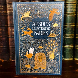 May include: A hardcover book titled "Aesop's Illustrated Fables" with a dark blue cover. The cover features gold illustrations of animals, including a cat, rabbit, lion, and peacock, along with the title text.