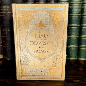 May include: A vintage copy of "The Iliad and the Odyssey" by Homer. The book has a beige cover with gold and blue illustrations of figures and patterns. It is displayed with other antique books, suggesting a collection.