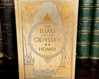 May include: A vintage book titled "The Iliad and the Odyssey" by Homer. The cover is beige with gold and blue decorative elements, including illustrations of figures and geometric patterns. The book is displayed among other antique books.