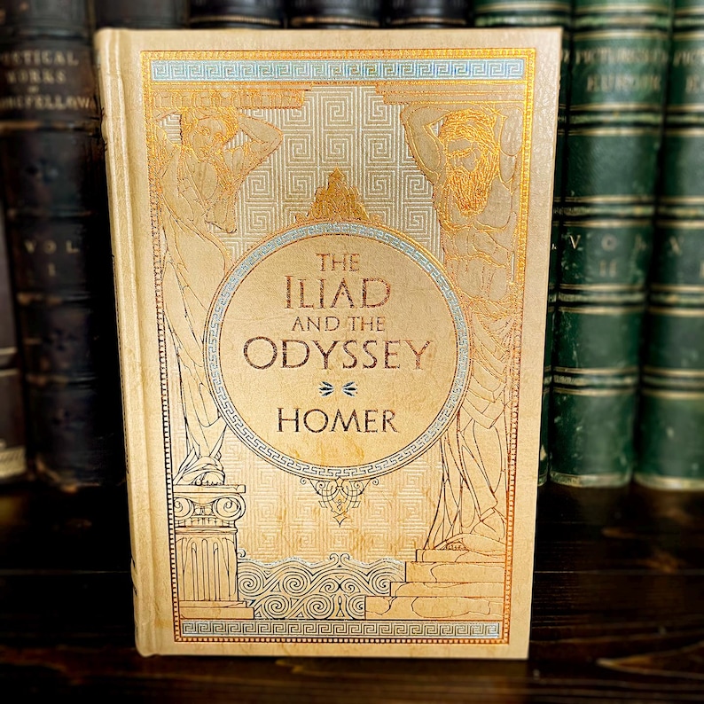 May include: A vintage copy of "The Iliad and the Odyssey" by Homer. The book has a beige cover with gold and blue illustrations of figures and patterns. It is displayed with other antique books, suggesting a collection.