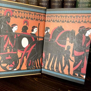 May include: A double-page spread from a book with illustrations of ancient Greek scenes. The artwork uses black, red, and orange, with a decorative border. The book is open on a dark wooden surface.