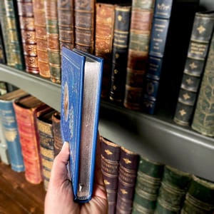 May include: A blue book with silver-gilded edges is held in front of a bookshelf filled with antique books. The cover of the book has a celestial design. The spines of the other books are in various colors, including brown, green, and blue.