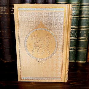 May include: Antique book with a tan cover featuring a gold geometric pattern and a circular illustration of two warriors. The spine is also tan. Other books are visible in the background.
