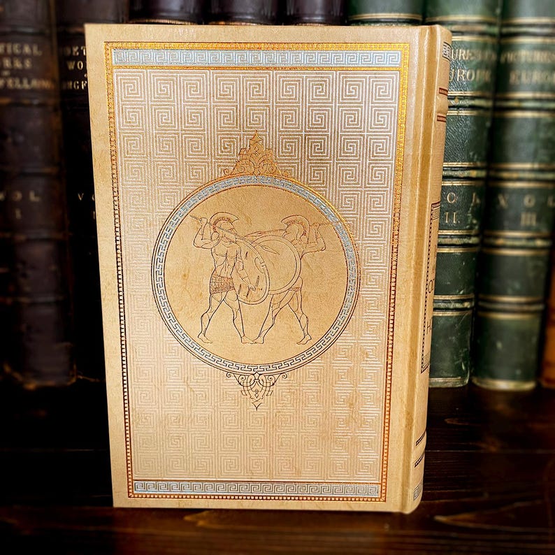 May include: Antique book with a tan cover featuring a gold geometric pattern and a circular illustration of two warriors. The spine is also tan. Other books are visible in the background.