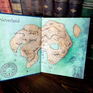 May include: Open book displaying a detailed map of Neverland, with the title at the top. The map features illustrations of Indian Village, Mermaid Lagoon, and Hook's Ship. The map is set against a teal background.