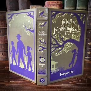 May include: A hardcover book titled "To Kill a Mockingbird" by Harper Lee. The book features a purple and gold design with illustrations of a family and a tree. The spine displays the title and author.