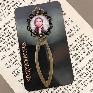 May include: A vintage-style bookmark featuring a circular image of a woman with brown braids, set within a decorative bronze-colored frame. The bookmark has a bronze-colored metal design and the text "STORYBOUNDERS" on a black background.