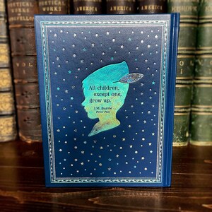 May include: A hardcover book with a dark blue cover featuring a silhouette of Peter Pan and the quote "All children, except one, grow up." The cover has a starry pattern and decorative border. The book is displayed in front of a collection of antique books.