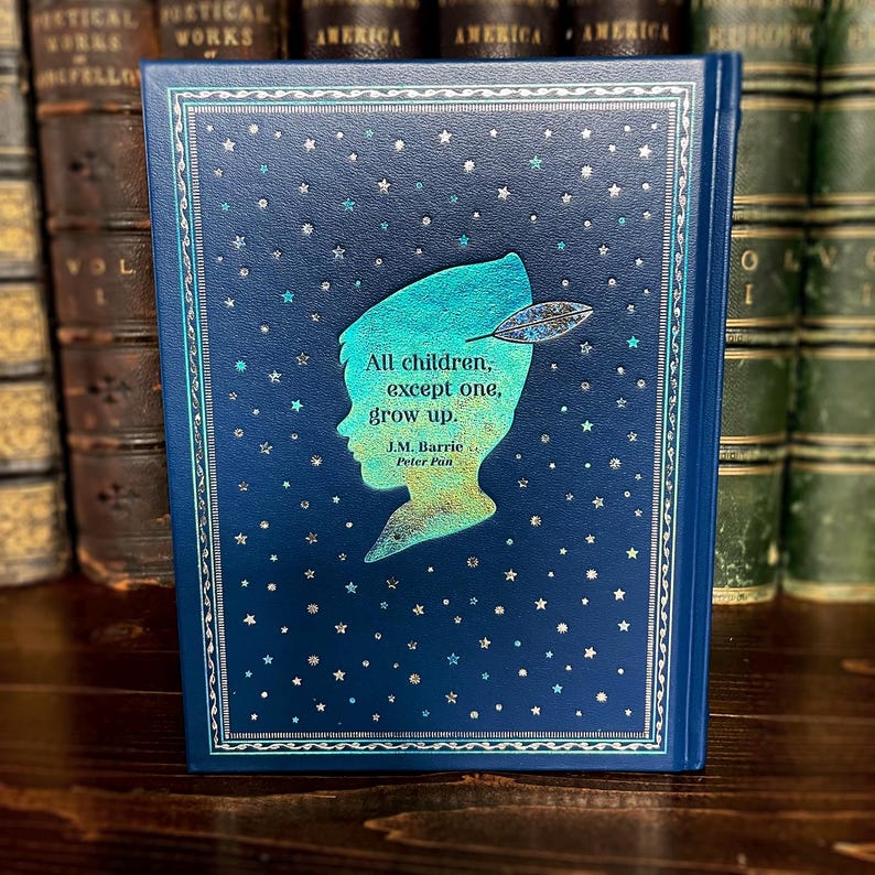 May include: A hardcover book with a dark blue cover featuring a silhouette of Peter Pan and the quote "All children, except one, grow up." The cover has a starry pattern and decorative border. The book is displayed in front of a collection of antique books.