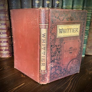 May include: Antique book titled "Whittier Illustrated" with a red cover and decorative floral and geometric patterns. The spine features the name "Whittier" vertically. The book is displayed on a dark wooden surface with other books in the background.