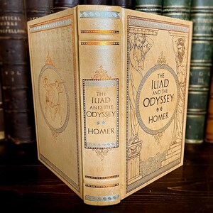 May include: A vintage hardcover book titled "The Iliad and the Odyssey" by Homer. The book has a cream-coloured cover with gold and blue decorative elements, including illustrations of figures and Greek key patterns. The spine displays the title and author.