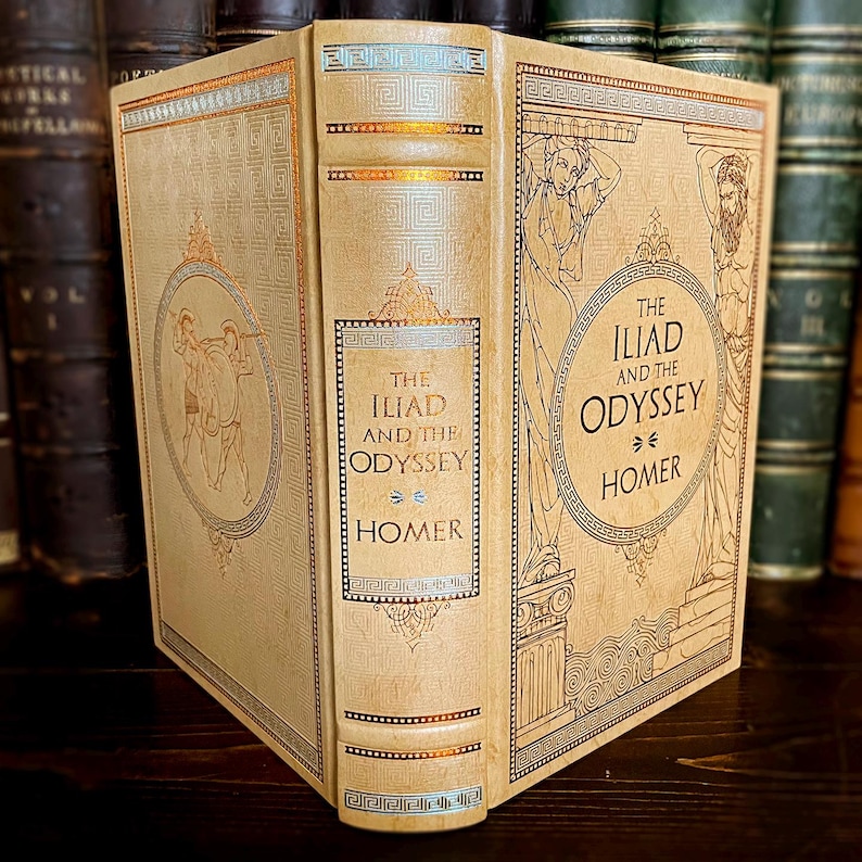 May include: A vintage hardcover book titled "The Iliad and the Odyssey" by Homer. The book has a cream-coloured cover with gold and blue decorative elements, including illustrations of figures and Greek key patterns. The spine displays the title and author.