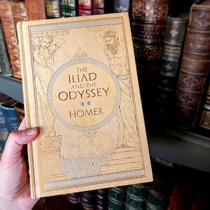 May include: A beige hardcover book titled "The Iliad and the Odyssey" by Homer. The cover features gold and blue illustrations of figures. The book is held in front of a shelf of antique books.