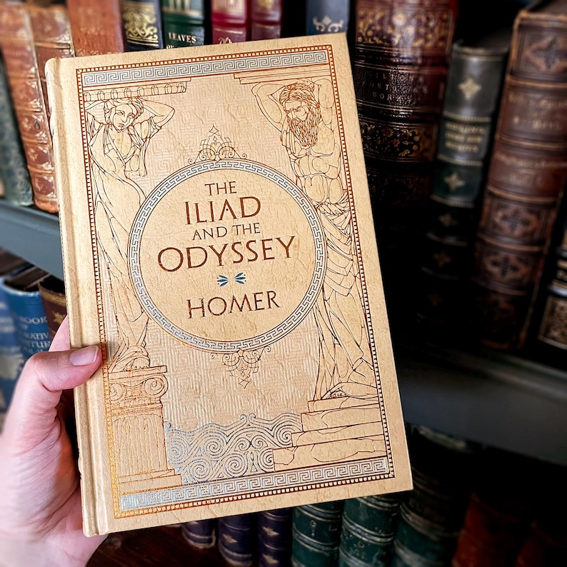 May include: A beige hardcover book titled "The Iliad and the Odyssey" by Homer. The cover features gold and blue illustrations of figures. The book is held in front of a shelf of antique books.
