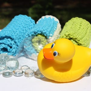 May include: A yellow rubber ducky toy with a bright orange beak and blue eyes. The ducky is surrounded by clear glass beads and three crocheted washcloths in blue, white, and green.