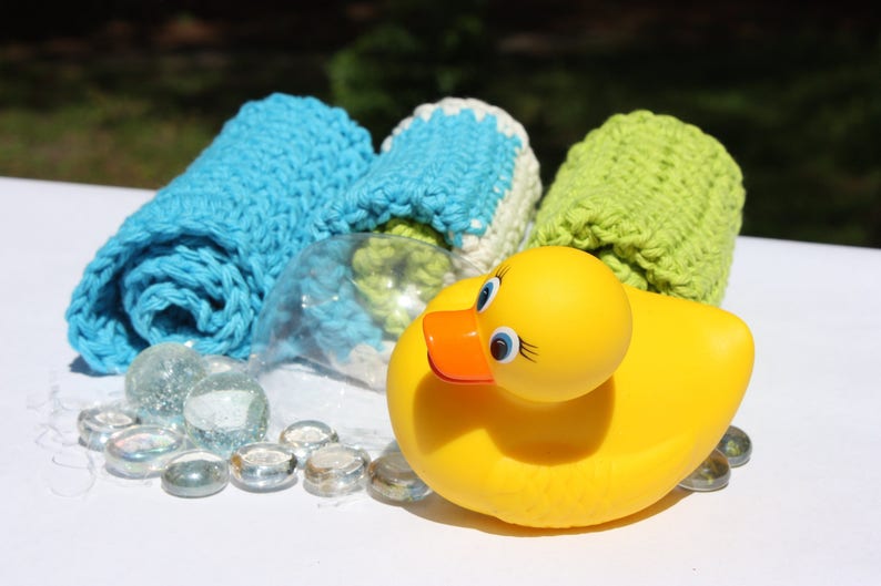 May include: A yellow rubber ducky toy with a bright orange beak and blue eyes. The ducky is surrounded by clear glass beads and three crocheted washcloths in blue, white, and green.