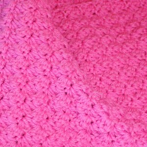 May include: Close-up of a vibrant pink crocheted blanket. The textured surface is created by the intricate stitch pattern, which gives the blanket a soft, plush appearance. The bright color and detailed texture make it visually appealing.