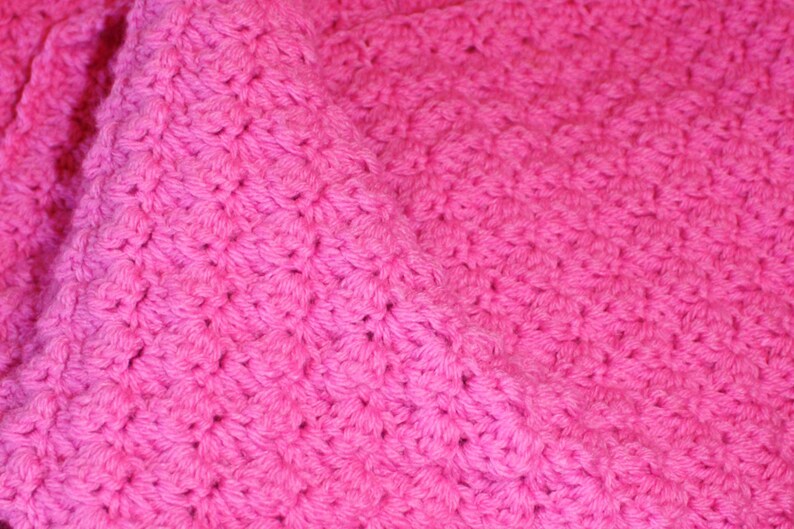 May include: Close-up of a vibrant pink crocheted blanket. The textured surface is created by the intricate stitch pattern, which gives the blanket a soft, plush appearance. The bright color and detailed texture make it visually appealing.