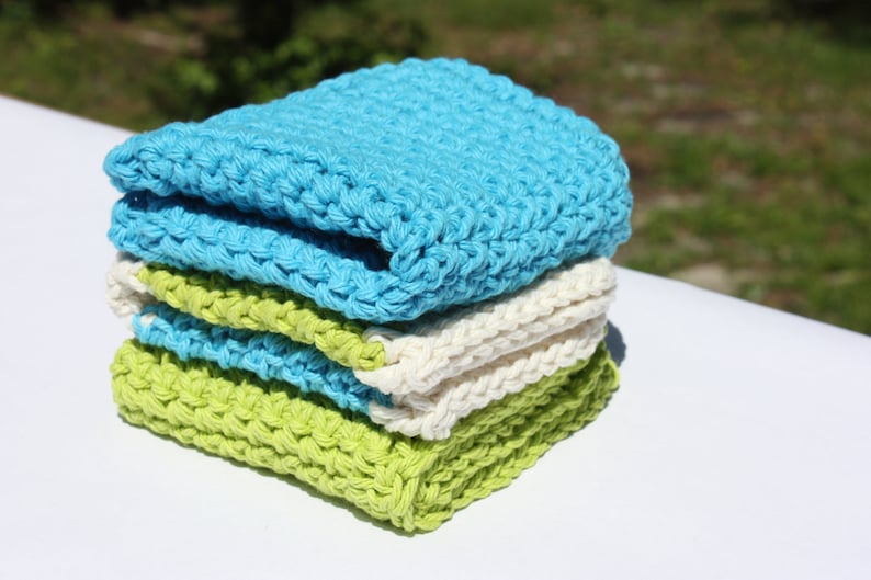 May include: A stack of four crocheted dishcloths in blue, white, and green. The dishcloths are made of cotton yarn and are perfect for cleaning dishes or wiping down surfaces.
