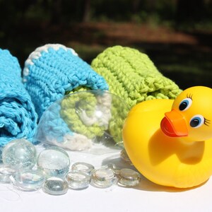 May include: Three crocheted washcloths in blue, green, and white, rolled up and placed on a white surface. A yellow rubber ducky is in the foreground.