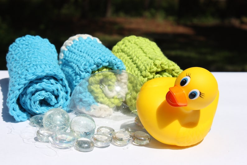 May include: Three crocheted washcloths in blue, green, and white, rolled up and placed on a white surface. A yellow rubber ducky is in the foreground.