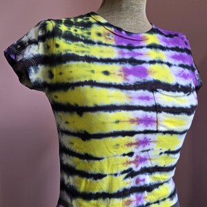 May include: A tie-dye t-shirt with a pocket, featuring horizontal stripes of yellow, black, and purple. The short-sleeved shirt is displayed on a mannequin. The shirt's design is a vibrant mix of colors.